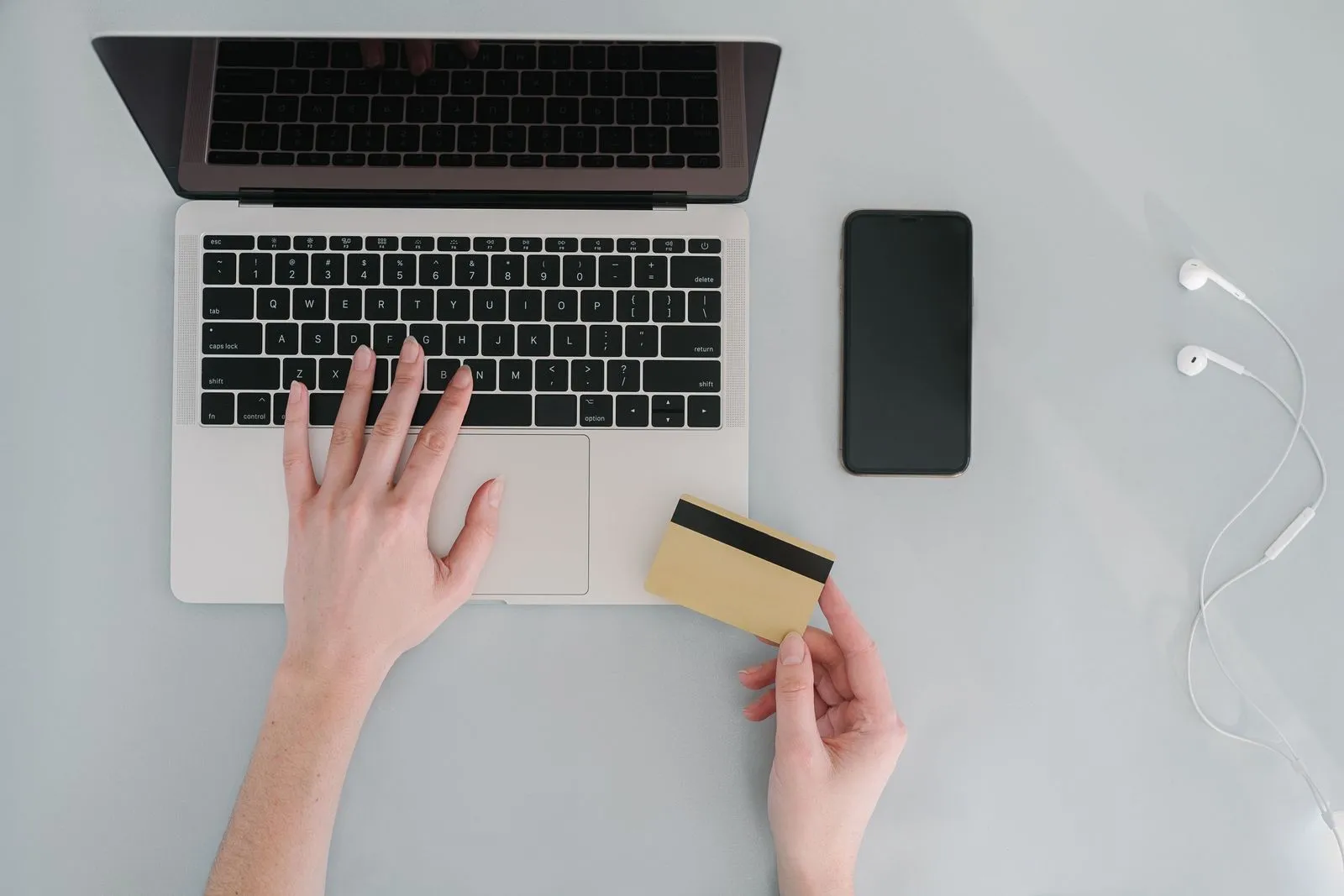 Mãos num notebook, uma delas segurando um cartão bancário dourado, com um celular sobre a mesa ao lado