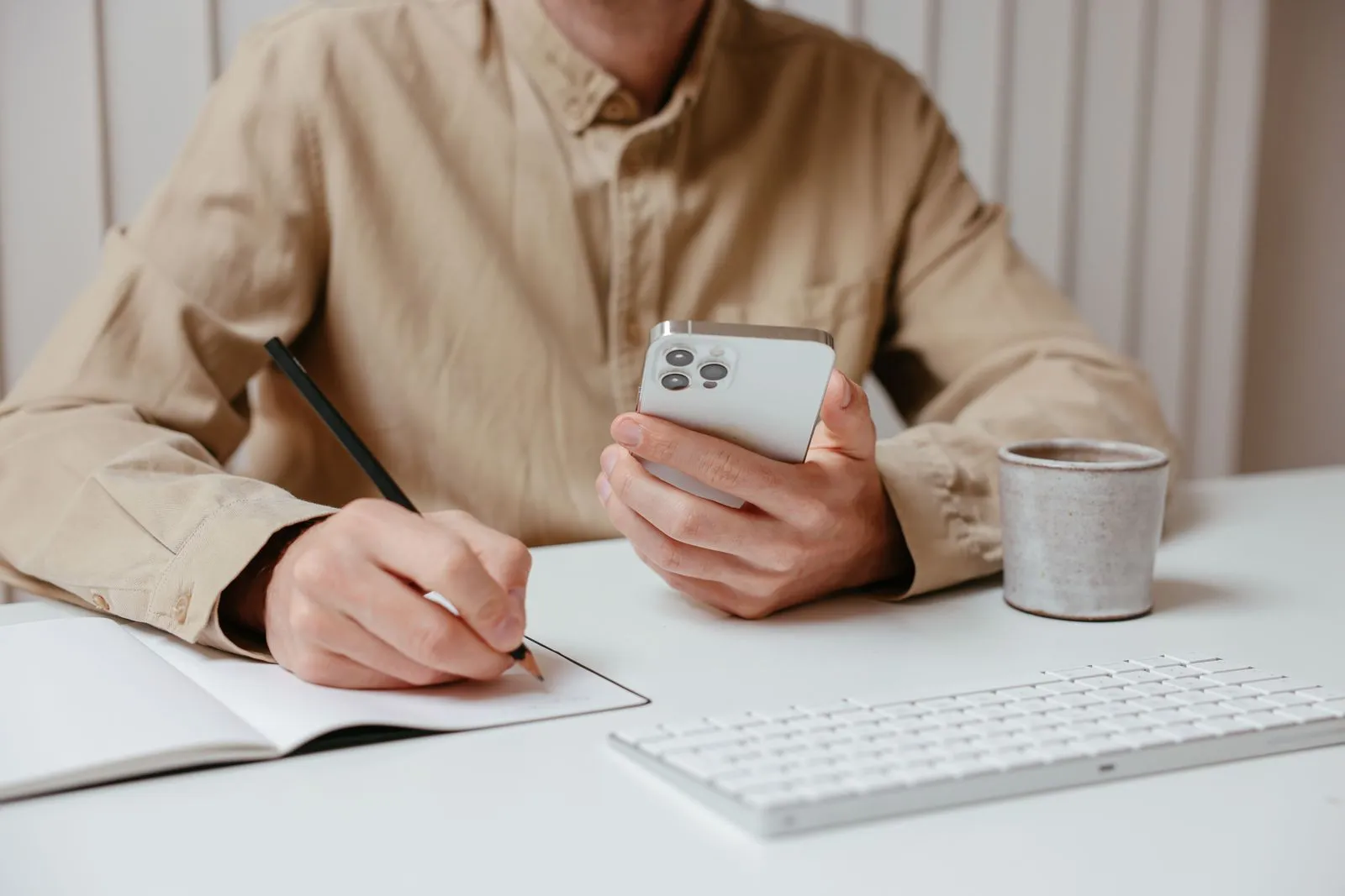 Uma pessoa escreve com lápis num caderno enquanto segura um celular, ao lado de um teclado e de uma caneca