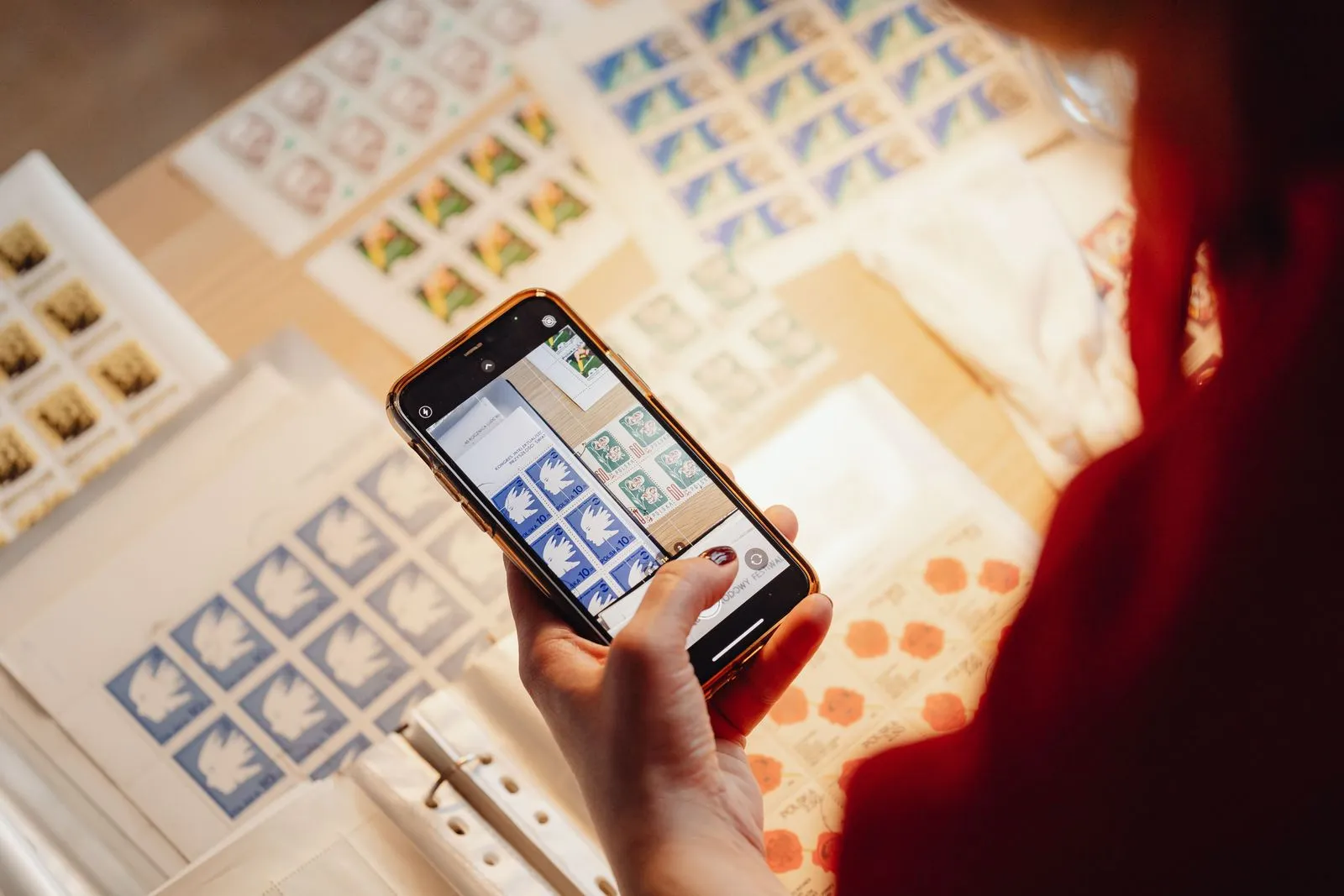 Una mano tiene uno smartphone sopra fogli di francobolli su un tavolo, sullo schermo è aperta l'app fotocamera