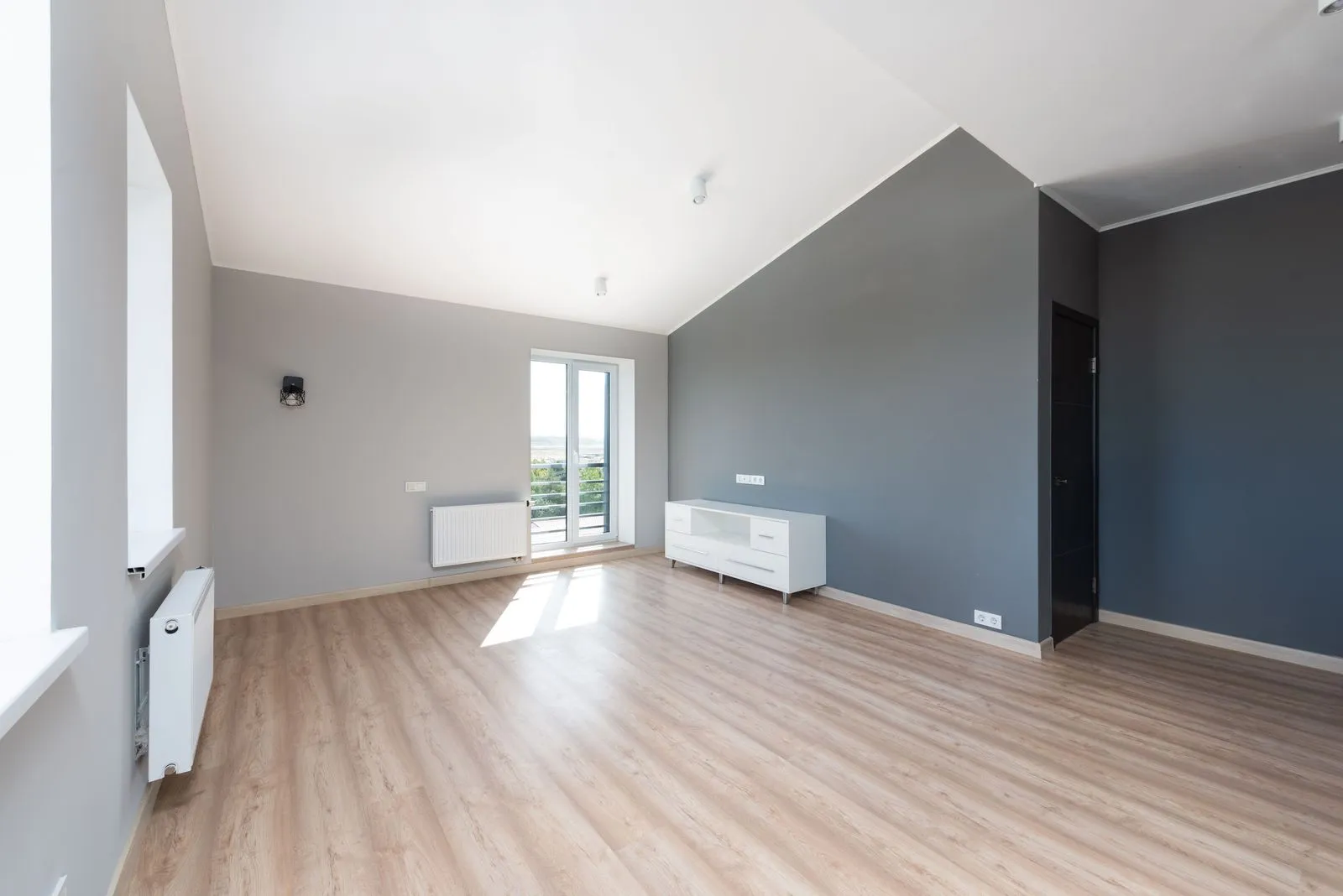 Appartamento luminoso e quasi vuoto con soffitto mansardato, pavimento in legno chiaro, pareti grigie e una porta finestra