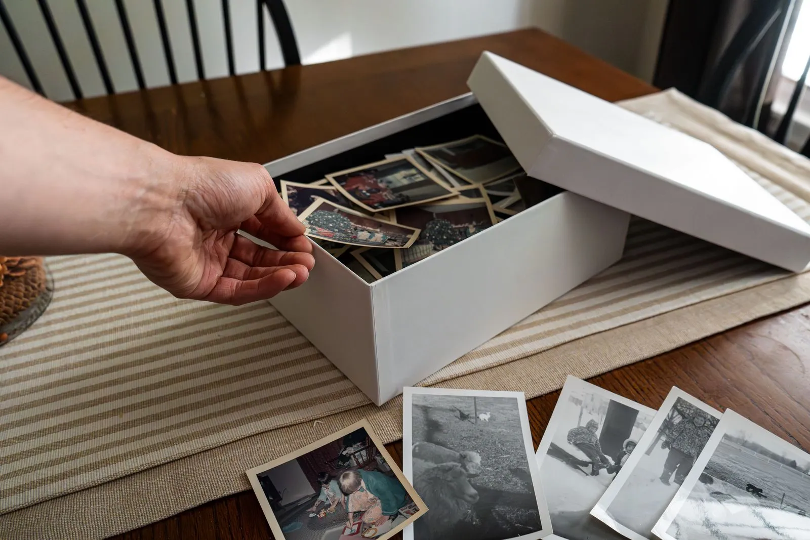 Mano che fruga in una scatola di vecchie foto di famiglia, altre stampe sparse sul tavolo